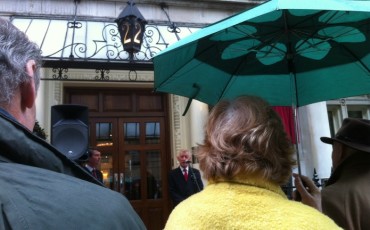 The historian Alan Judd speaking on the mic, with Tim Laurence, chair of the Blue Plaque Trust, standing near the loud speaker.