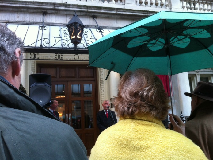 The historian Alan Judd speaking on the mic, with Tim Laurence, chair of the Blue Plaque Trust, standing near the loud speaker.