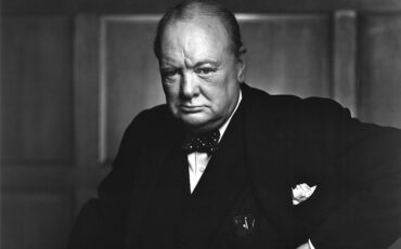 Churchill, aged 67, wearing a suit, standing and holding into the back of a chair. Photo Credit: © Public Domain via Wikimedia Commons.