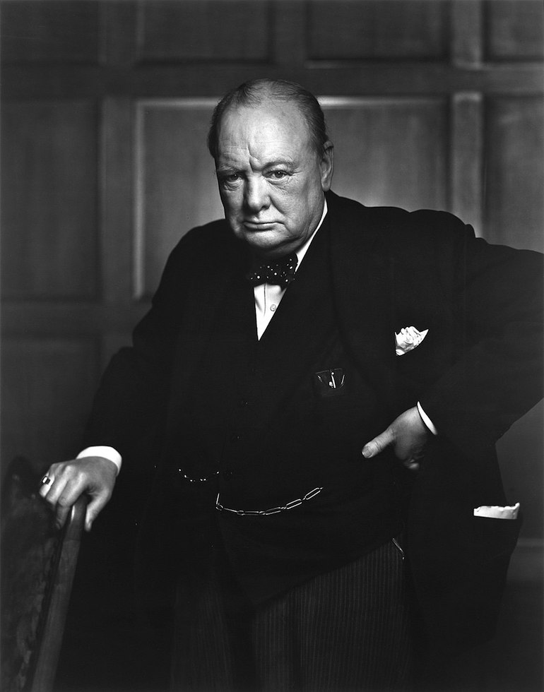 Churchill, aged 67, wearing a suit, standing and holding into the back of a chair. Photo Credit: © Public Domain via Wikimedia Commons. 